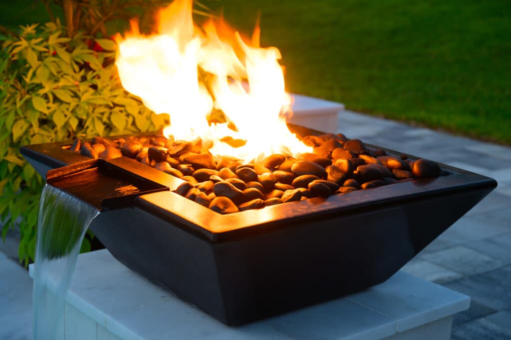 Black fire water bowl with flickering flames, set against the backdrop of the pool and greenery.