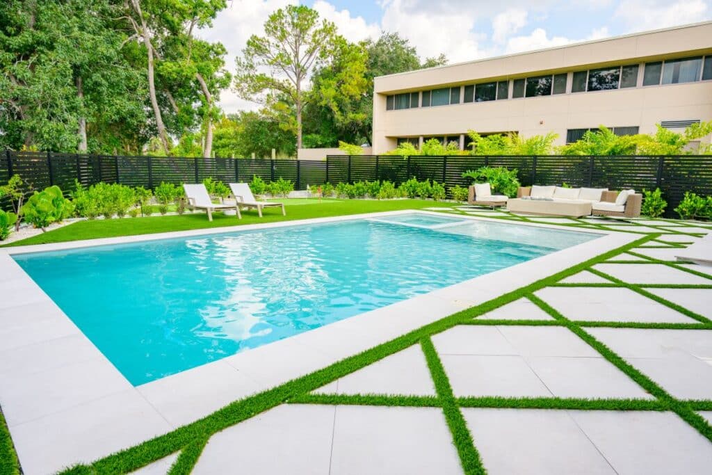 Sunlit backyard pool with a lawn area and geometric pavers.