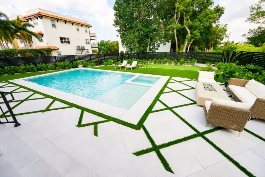 Wide shot of a poolside seating area with light stone pavers and green grass accents.