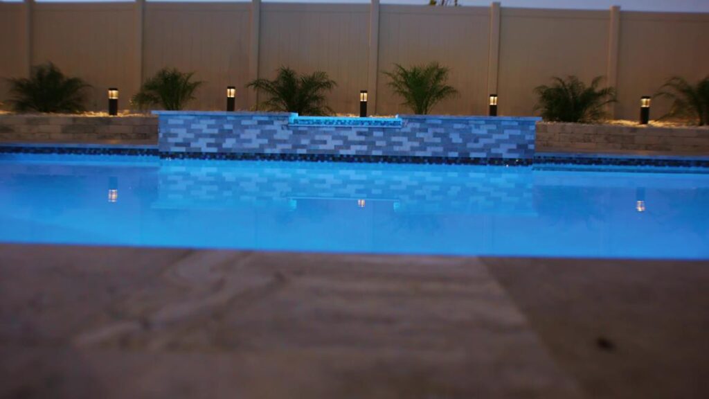 A backyard pool with a stylish water feature and well-lit landscaping, highlighting a polished tile pool deck.