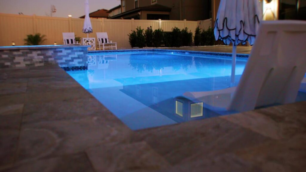 A nighttime view of a pool with integrated pool lights and reflective water, showcasing a luxurious backyard setting.