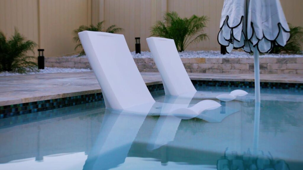 A pair of poolside lounge chairs resting in the water on a sunshelf, shaded by an umbrella.