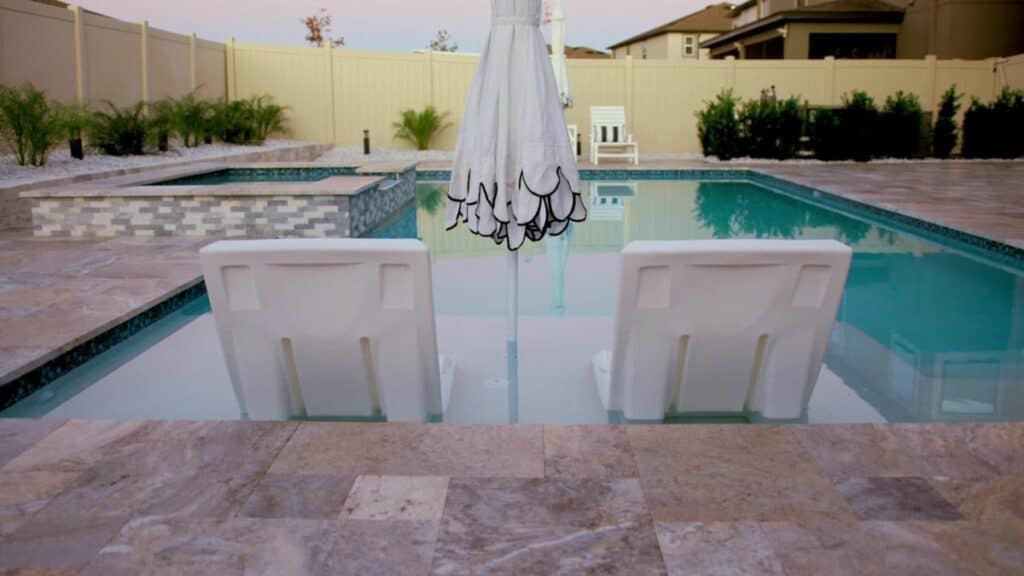 Lounge chairs on a pool sun shelf under an elegant umbrella, surrounded by travertine decking and soft landscaping.